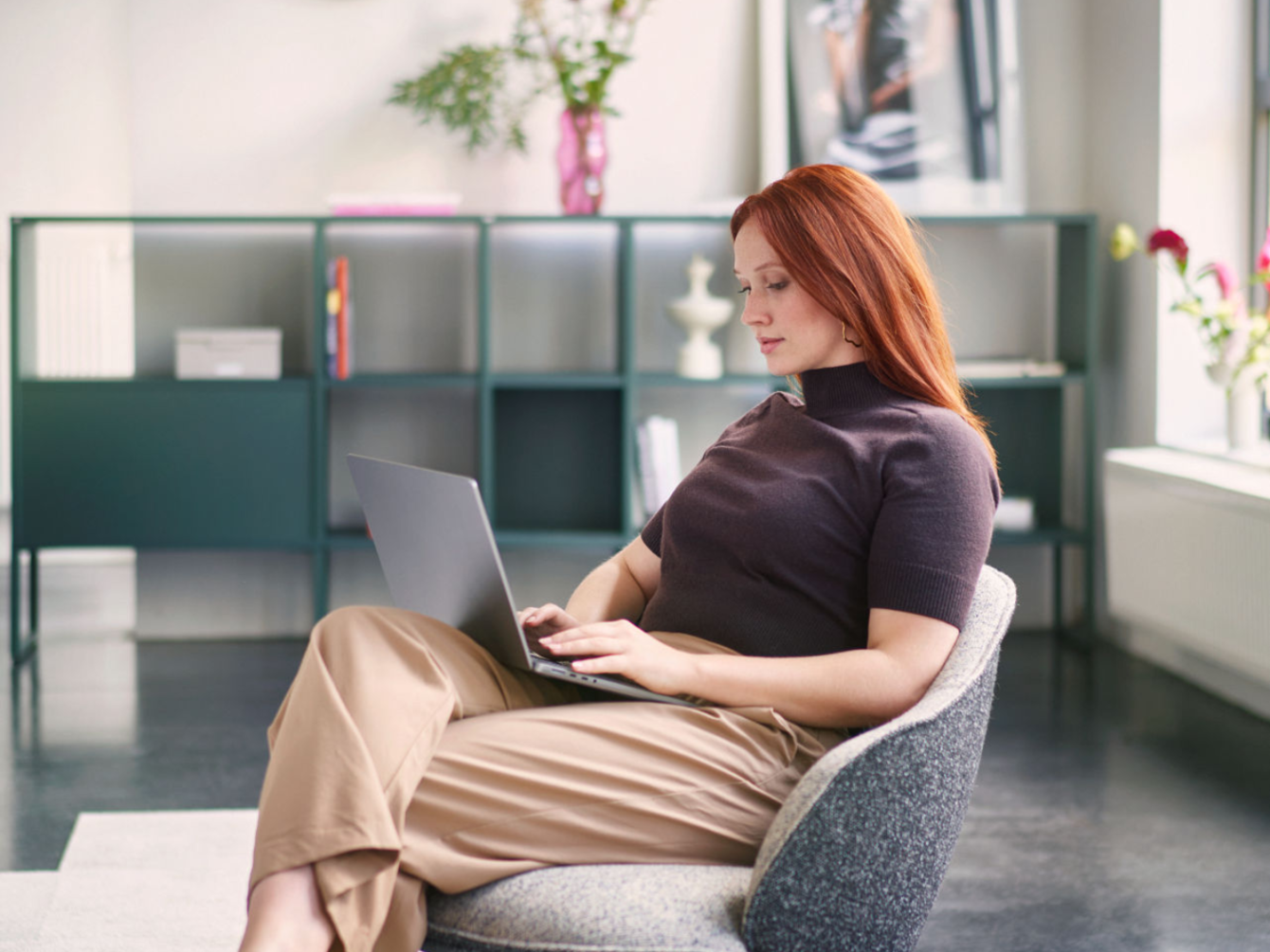 Mujer pelirroja sentada relajada en un sillón gris trabajando con un portátil en una habitación moderna con estanterías y plantas al fondo