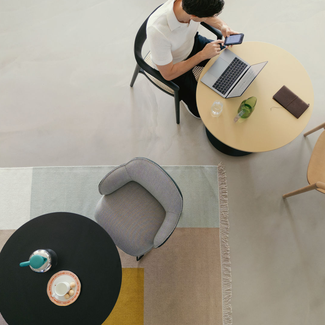 Bird's-eye view of a person sitting at a round table with a laptop, smartphone, glass, and book, surrounded by chairs and a geometric patterned rug 