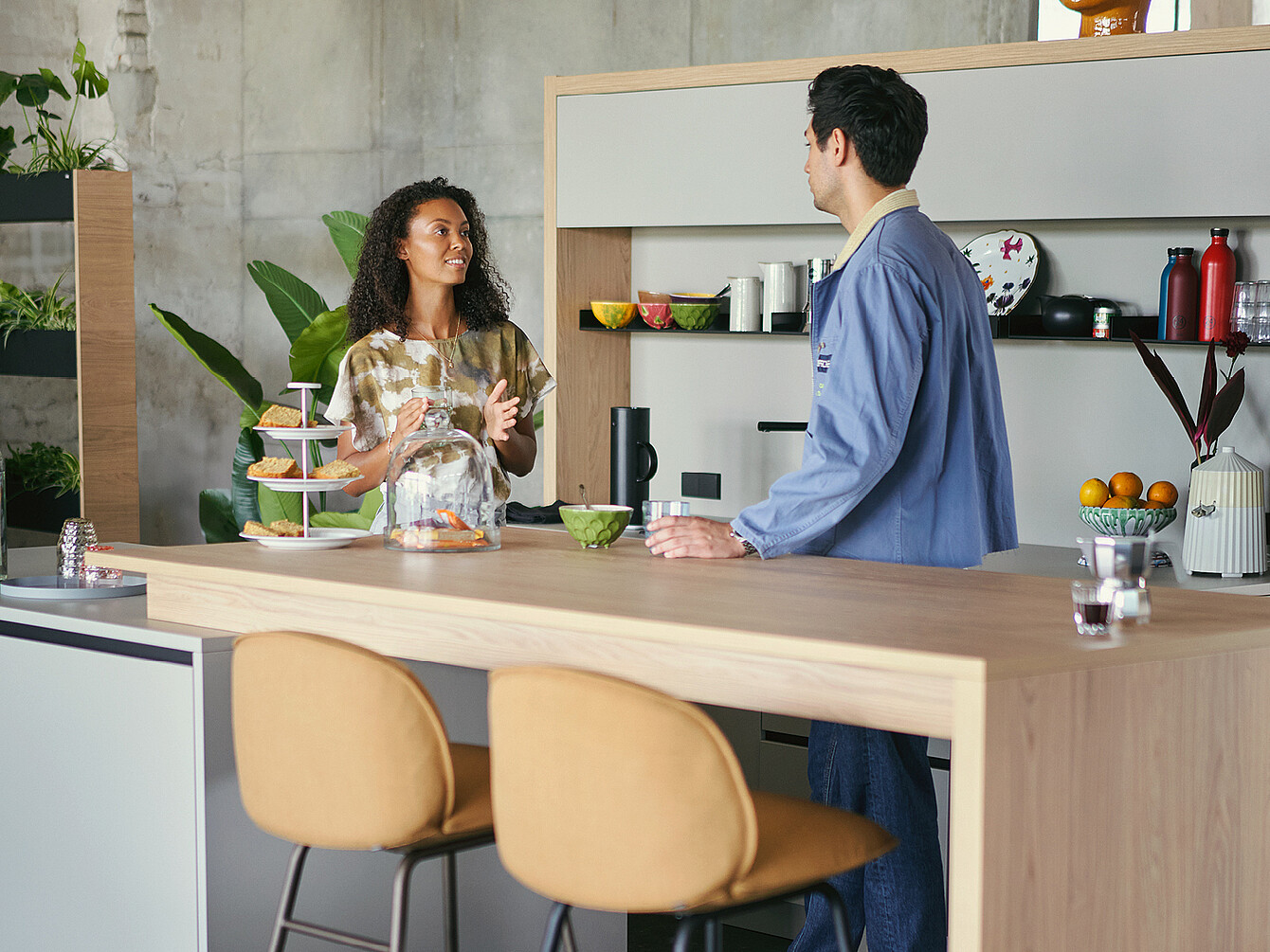 Dos empleados conversan en la barra de la cocina de una oficina moderna con aperitivos y bebidas a la vista, lo que pone de relieve la interacción intencionada en la oficina.