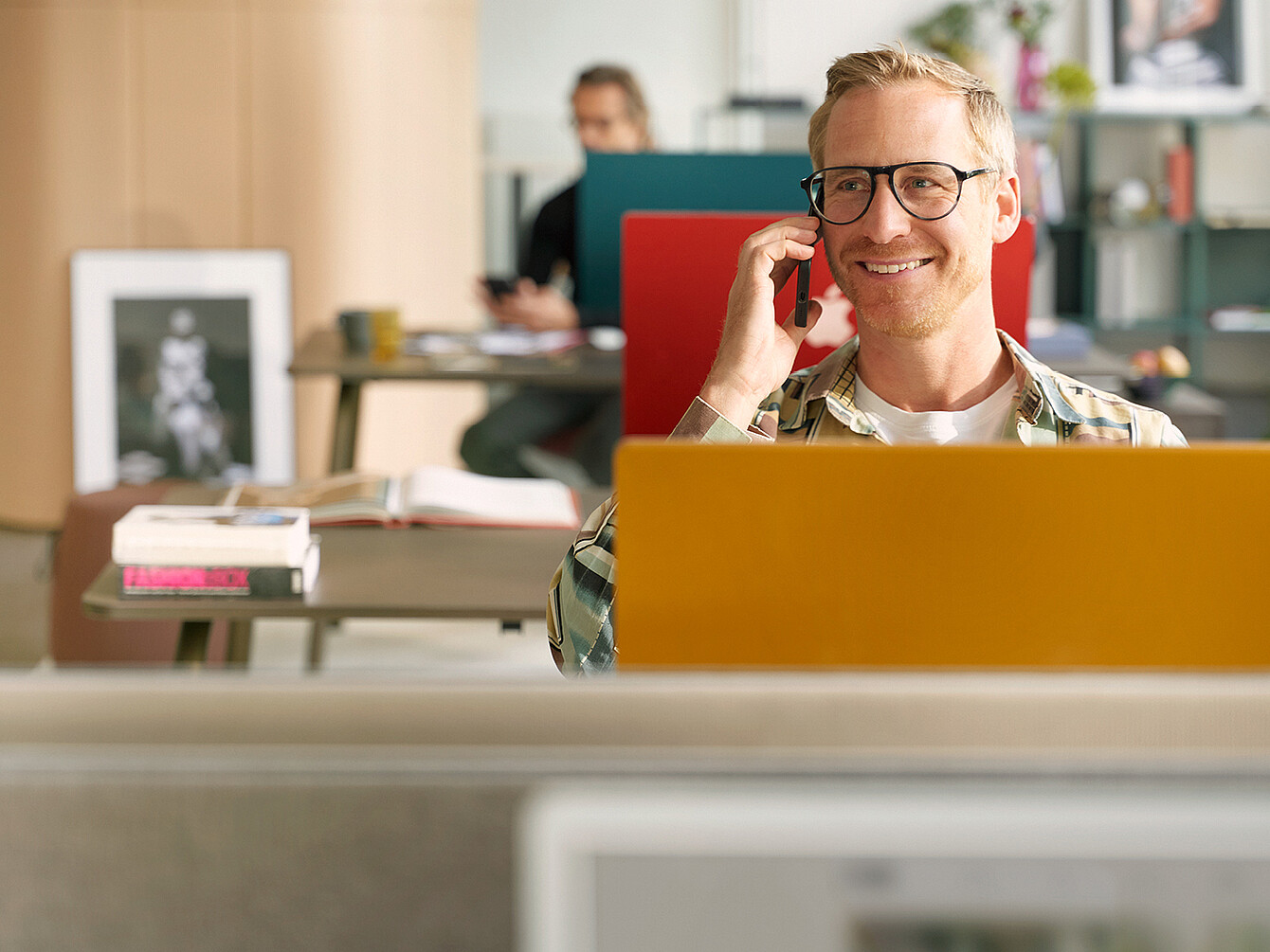 Hombre con gafas sentado detrás de un portátil amarillo en un espacio de oficina hablando por teléfono con una persona que trabaja en un escritorio al fondo