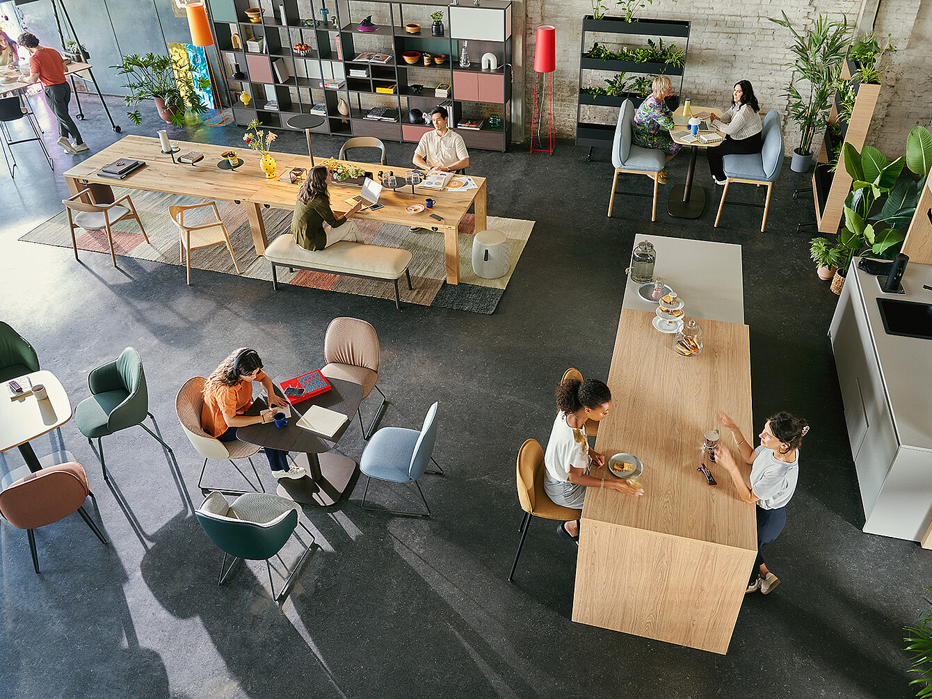 Offener Arbeitsbereich mit mehreren Personen, die an Tischen arbeiten und sich unterhalten, umgeben von Pflanzen und Regalen