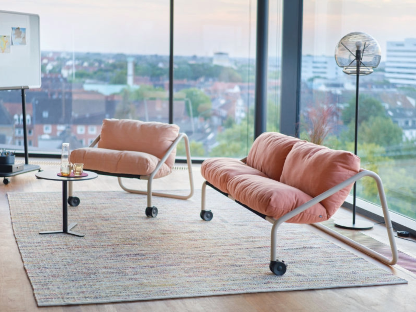 Modernes Sofa und Sessel mit Metallgestell und Rollen auf einem Teppich vor bodentiefen Fenstern mit Stadtblick