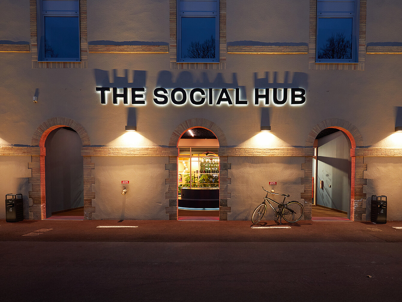 Façade d’un bâtiment avec trois entrées en arc éclairées au crépuscule, un vélo garé près d’une entrée et une enseigne « THE SOCIAL HUB » au-dessus de l’arc central. 