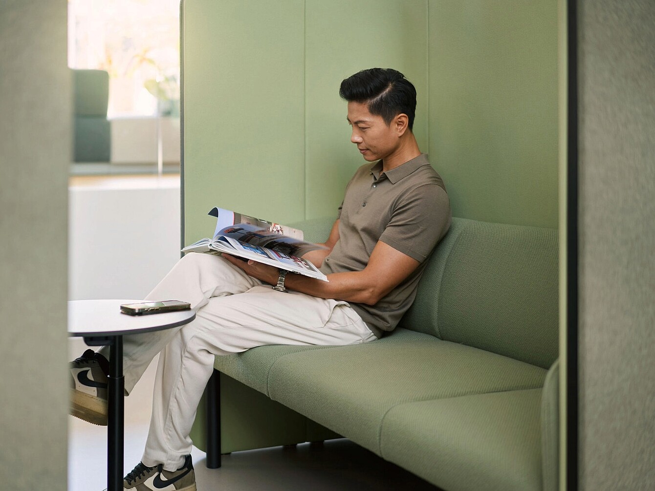 Hombre sentado en un banco tapizado en verde en una solución de sala se:hive leyendo una revista con un smartphone en una pequeña mesa redonda cercana