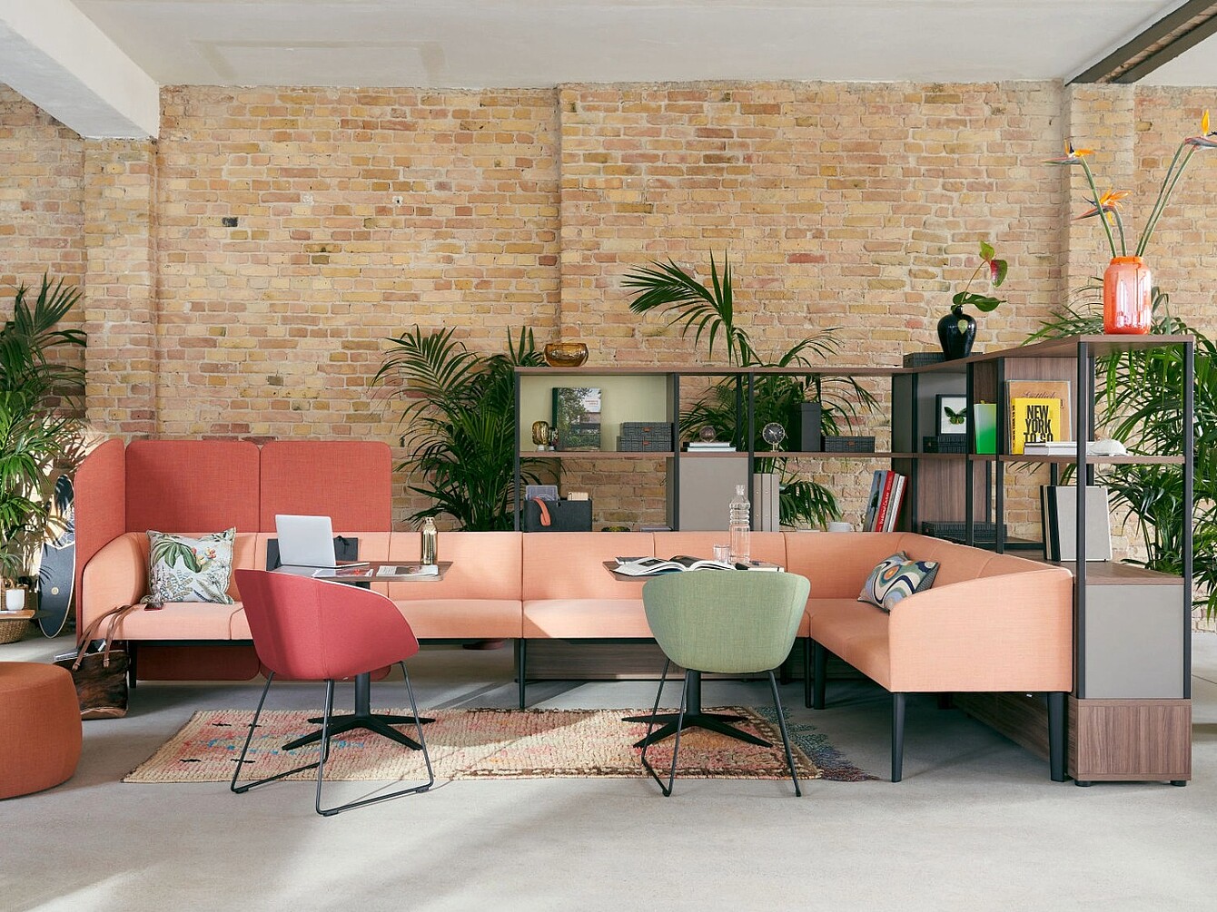 Area lounge aperta con un divano componibile color pesca, due sedie moderne in rosso e verde, tavolini bassi, piante e una scaffalatura contro un muro di mattoni