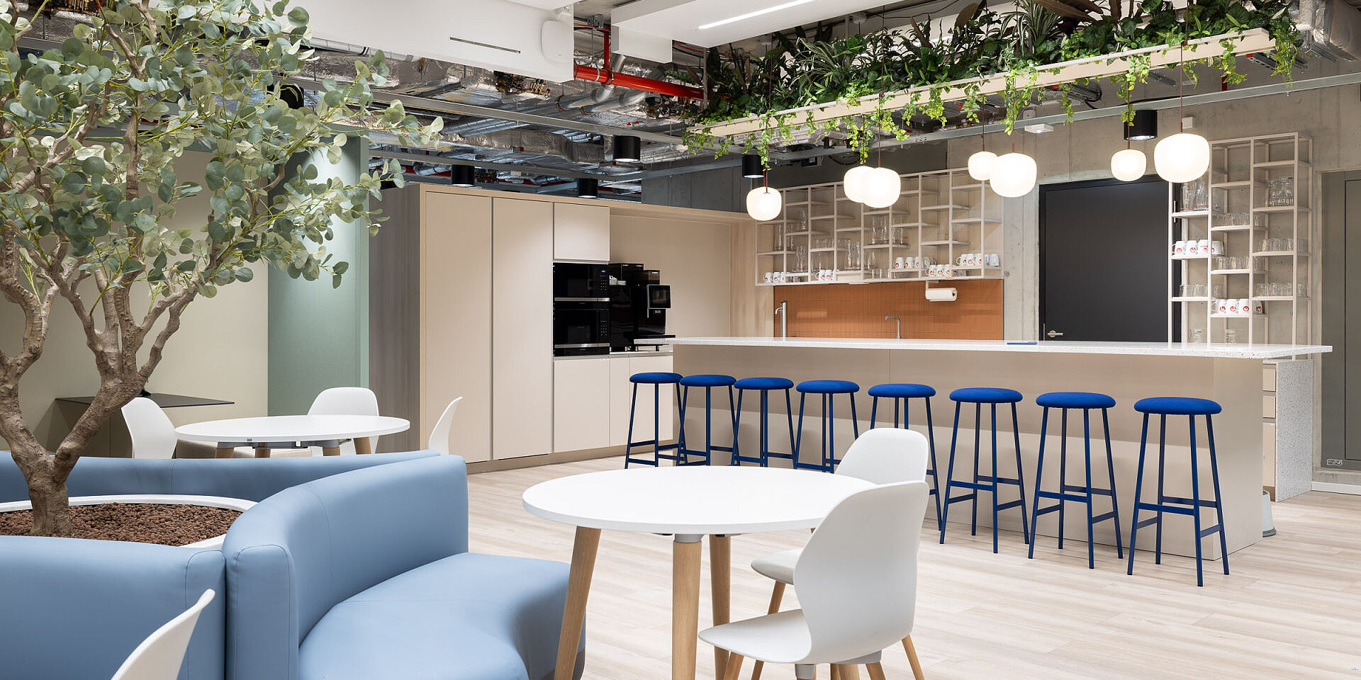 Modern bistro area at Veolia with se:spot designer chairs in white and blue bar stools at se:assist tables, surrounded by plants and bright lighting. 