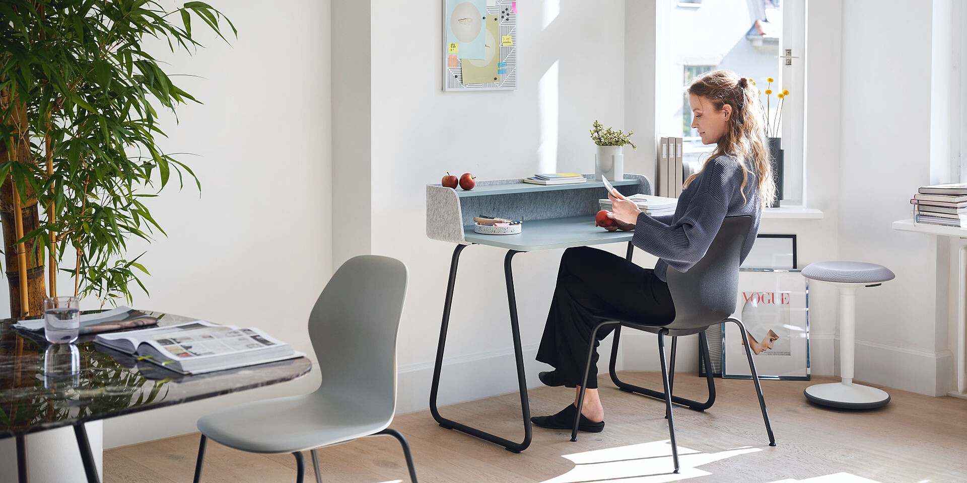 Woman sitting at a modern desk with black legs and a grey surface, reading a document in a bright, minimalist workspace with plants and large windows.