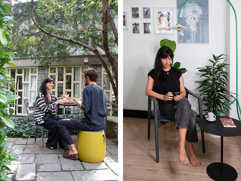Two people are sitting outside on yellow stools at a small table in the garden. One person is sitting barefoot indoors on a chair next to a black side table with a cup and a book, surrounded by modern wall art and a green plant.