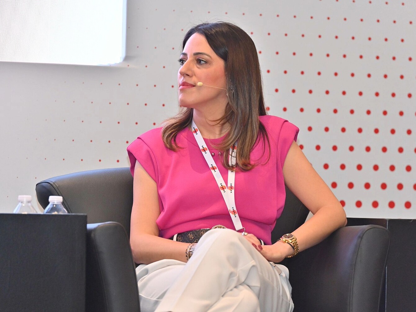 Donna con top rosa senza maniche e pantaloni chiari seduta a gambe incrociate su una poltrona nera davanti a una parete chiara con un motivo a pois rossi