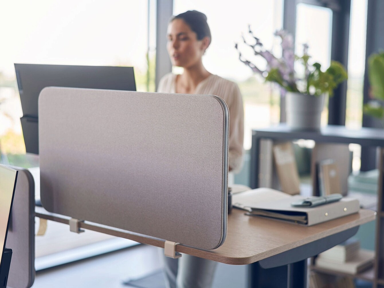 De afbeelding toont een moderne kantoorwerkplek met een bureau en een akoestische scheidingswand, die meer privacy biedt en de concentratie helpt verbeteren. Op de achtergrond werkt iemand achter een computer, terwijl planten en open rekken zorgen voor een aangename en professionele werkomgeving. De indeling ondersteunt geconcentreerd werken binnen een open kantoorindeling.