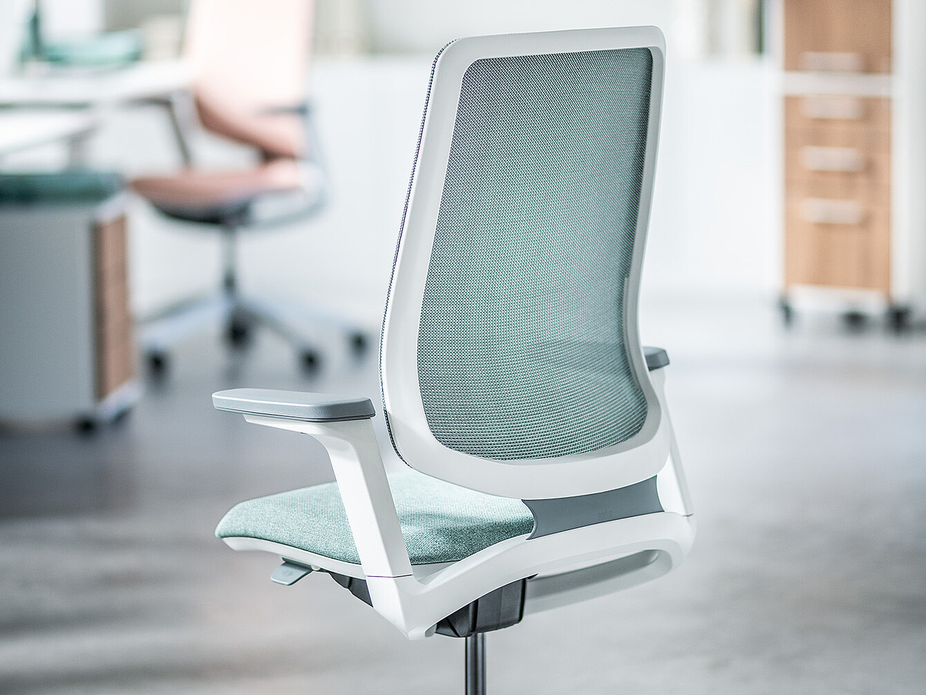 Silla de oficina ergonómica de color verde claro con respaldo de malla y estructura blanca en un entorno de oficina moderno