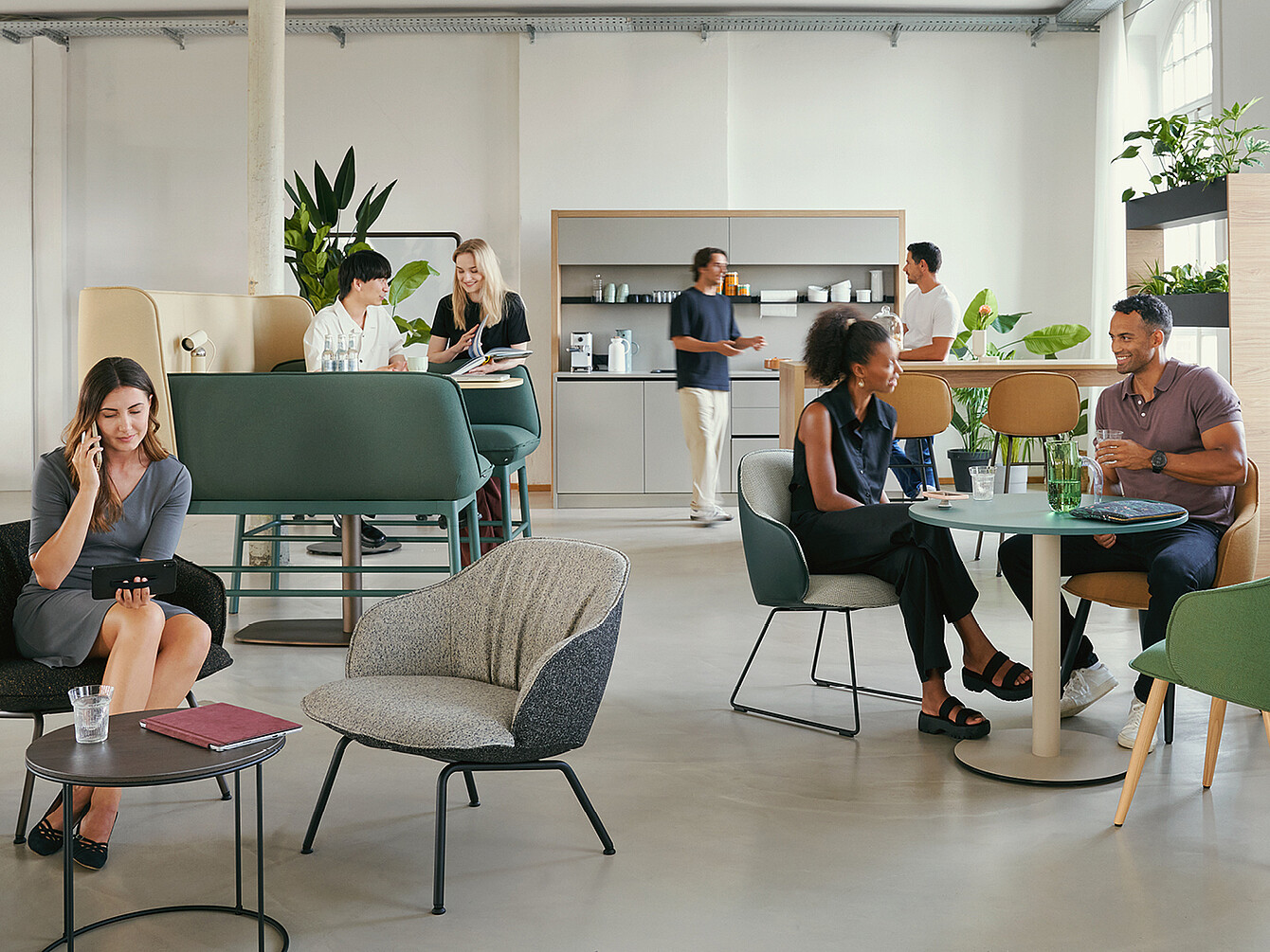 Un moderno Work Café con varie disposizioni dei posti a sedere. Le persone sono impegnate in conversazioni, alcune utilizzano il telefono. Sono visibili piante verdi e sullo sfondo si intravede un angolo cottura.