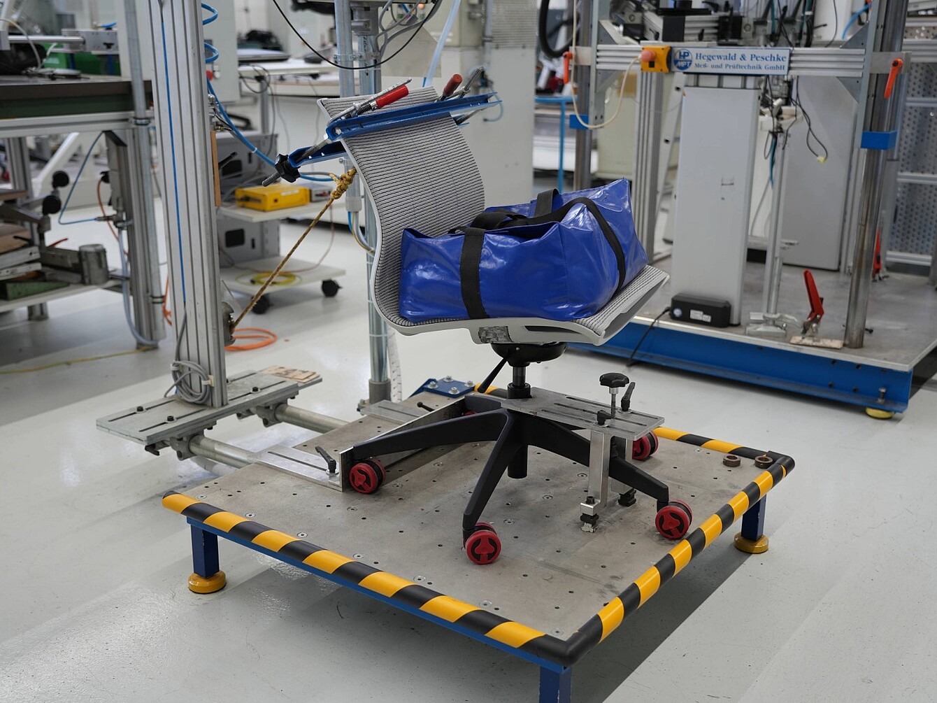 Silla de oficina con respaldo de malla y bolsa azul sobre una plataforma de ensayo con bordes a rayas amarillas y negras en un entorno de laboratorio