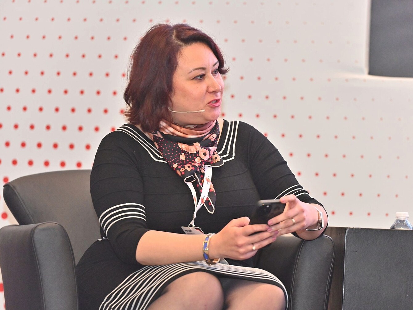 Donna seduta in abito nero a righe bianche e sciarpa fantasia che tiene in mano uno smartphone durante una conferenza