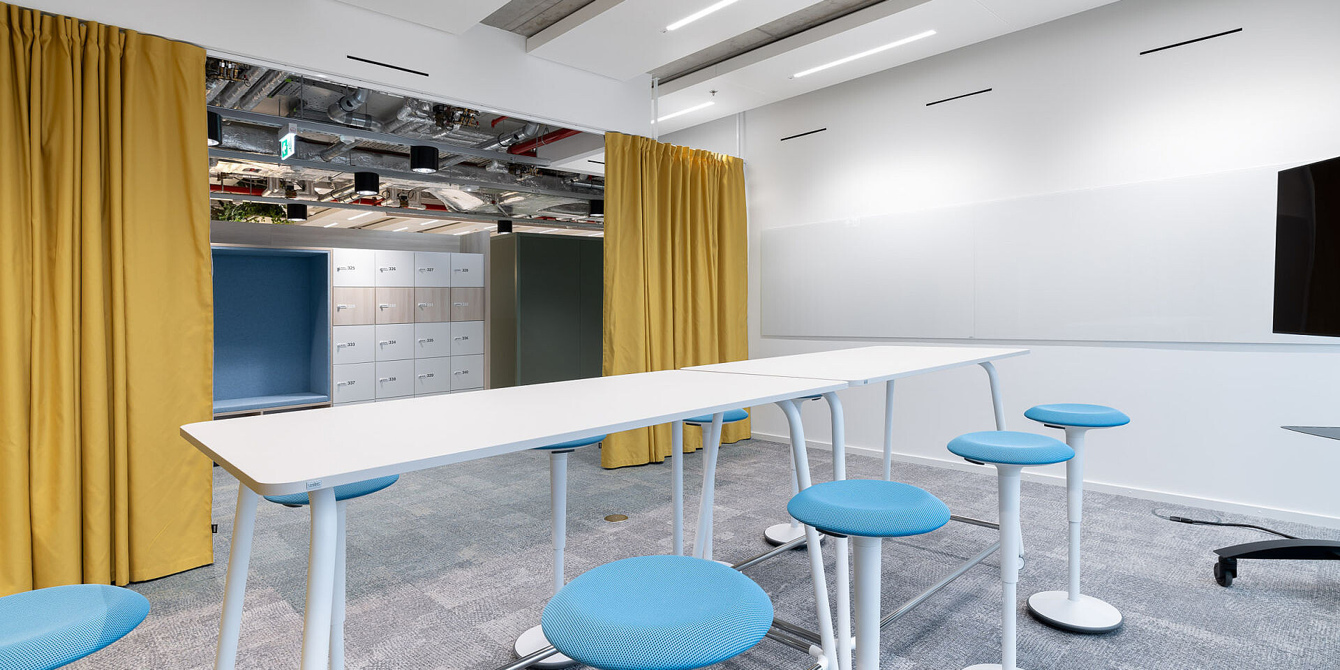 Modern meeting room with white se:lab high desk workshop table surrounded by blue se:fit standing seats and se:dot stools, yellow curtains and white walls. 