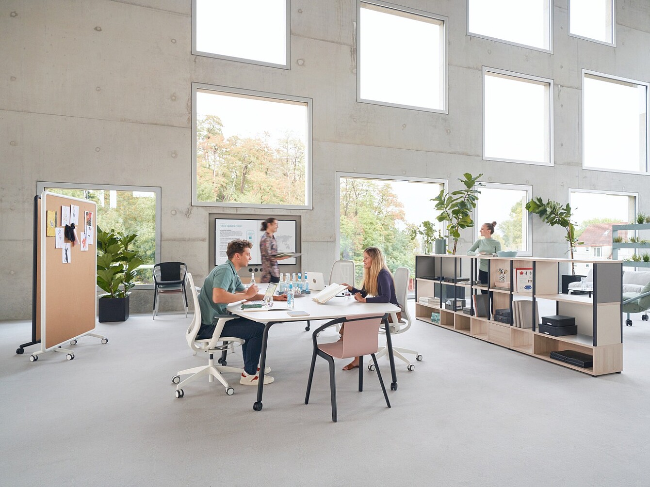 Due persone sedute a un tavolo che lavorano con documenti e computer portatili in un ufficio luminoso con ampie finestre quadrate e una scaffalatura