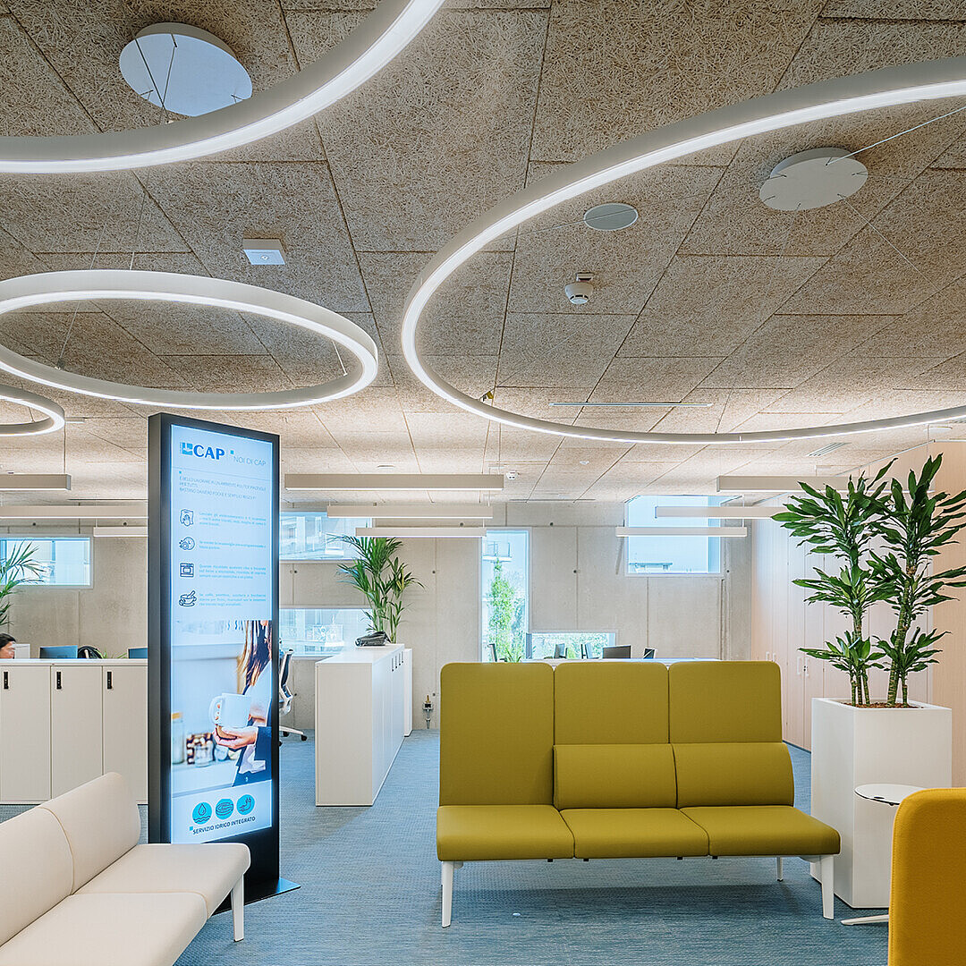 Offenes Büro mit modernen Sitzbänken, großen Pflanzen in weißen Töpfen und einem digitalen Informationsdisplay unter kreisförmigen Deckenleuchten