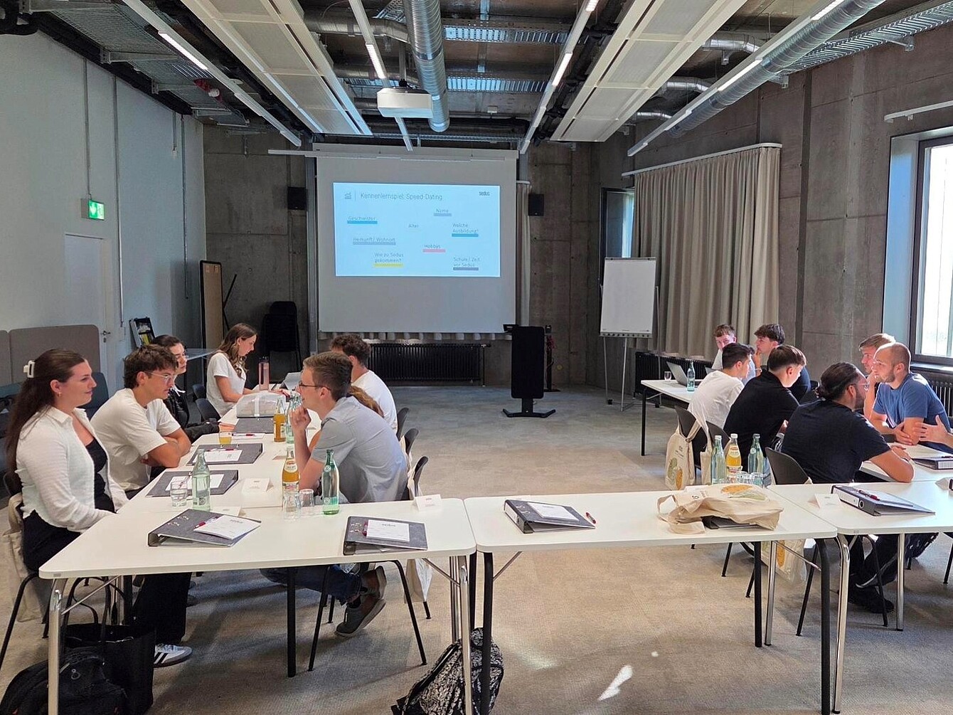Mehrere junge Menschen sitzen paarweise an weißen Tischen in einem hellen, industriell gestalteten Raum mit sichtbaren Rohren und einer Projektionsleinwand im Hintergrund