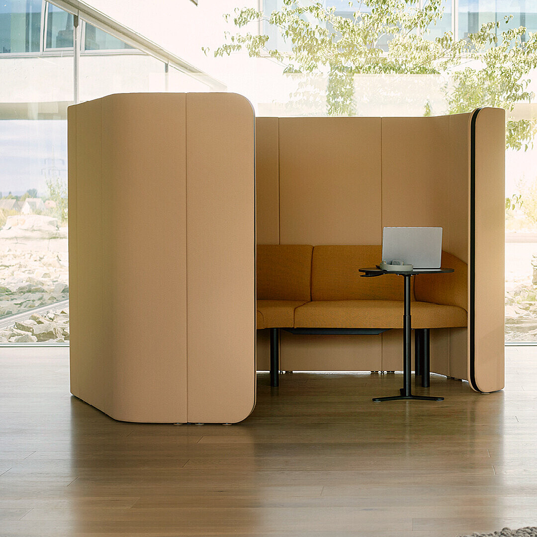 Moderner, offener Raum mit einem beige-farbenen, halb geschlossenen Sitzbereich, einem kleinen schwarzen Tisch mit Laptop, einem grauen Sessel, einem Garderobenständer und einem runden Tisch mit zwei Tassen vor großen Fenstern.
