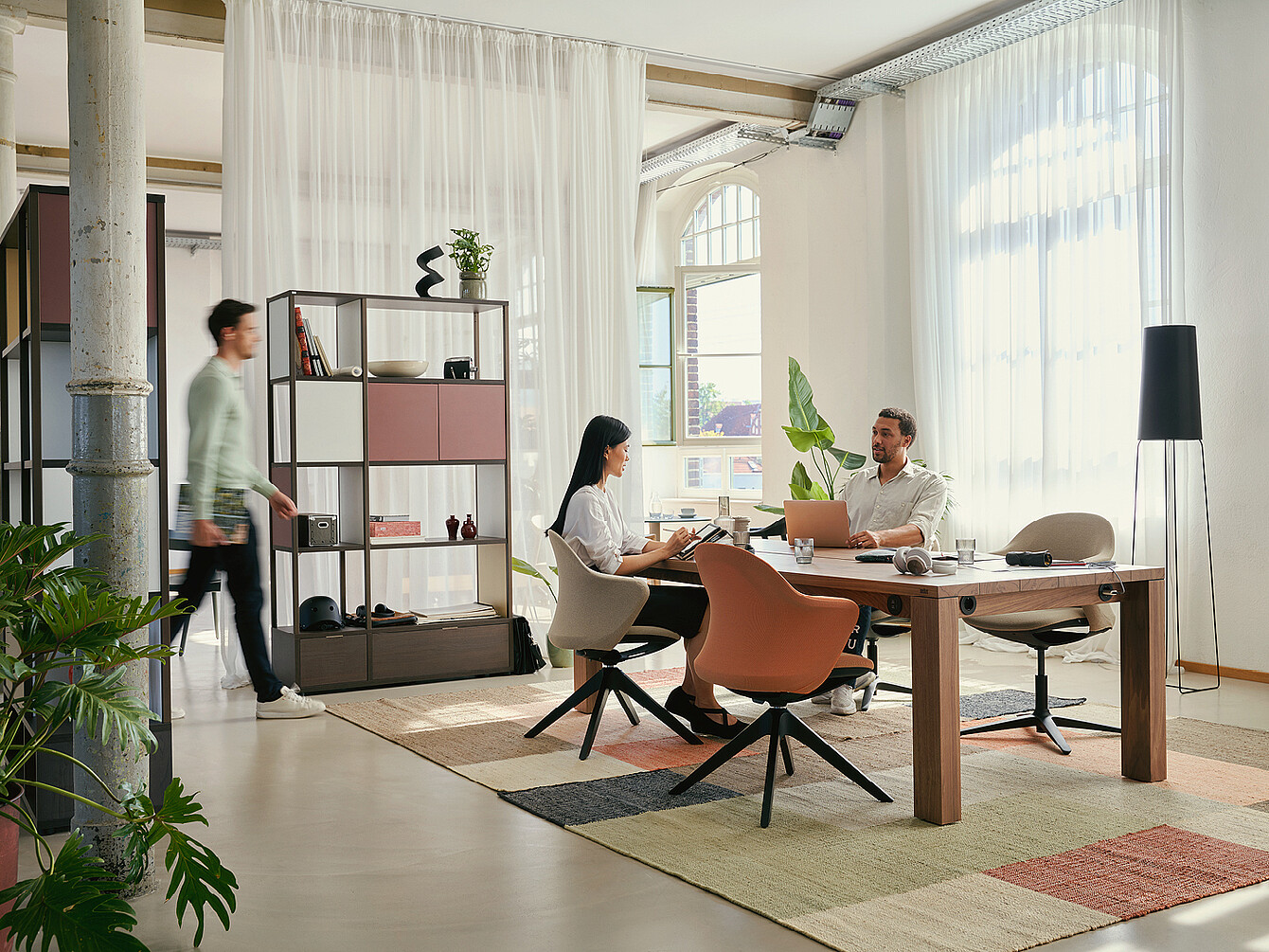 Moderno spazio di lavoro ibrido e flessibile con due persone sedute a un tavolo di legno che utilizzano laptop e tablet, circondate da piante, scaffali e ampie finestre con tende trasparenti, che enfatizzano la flessibilità e la comunità.
