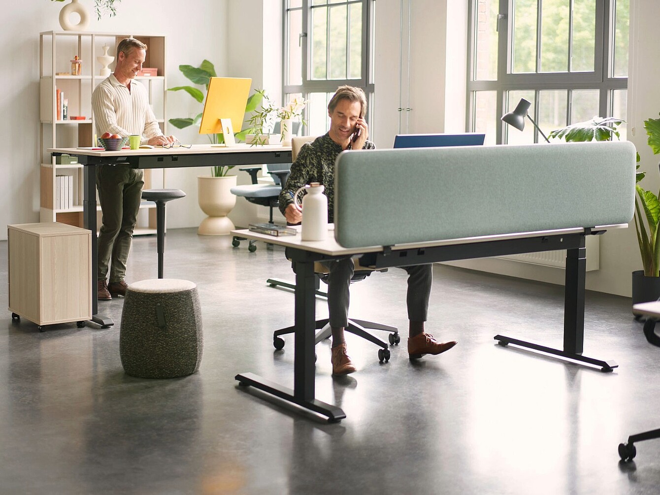 Espacio de oficina moderno con dos personas trabajando en escritorios separados por una pantalla de privacidad azul claro, con sillas ergonómicas, un escritorio de pie y luz natural procedente de grandes ventanas