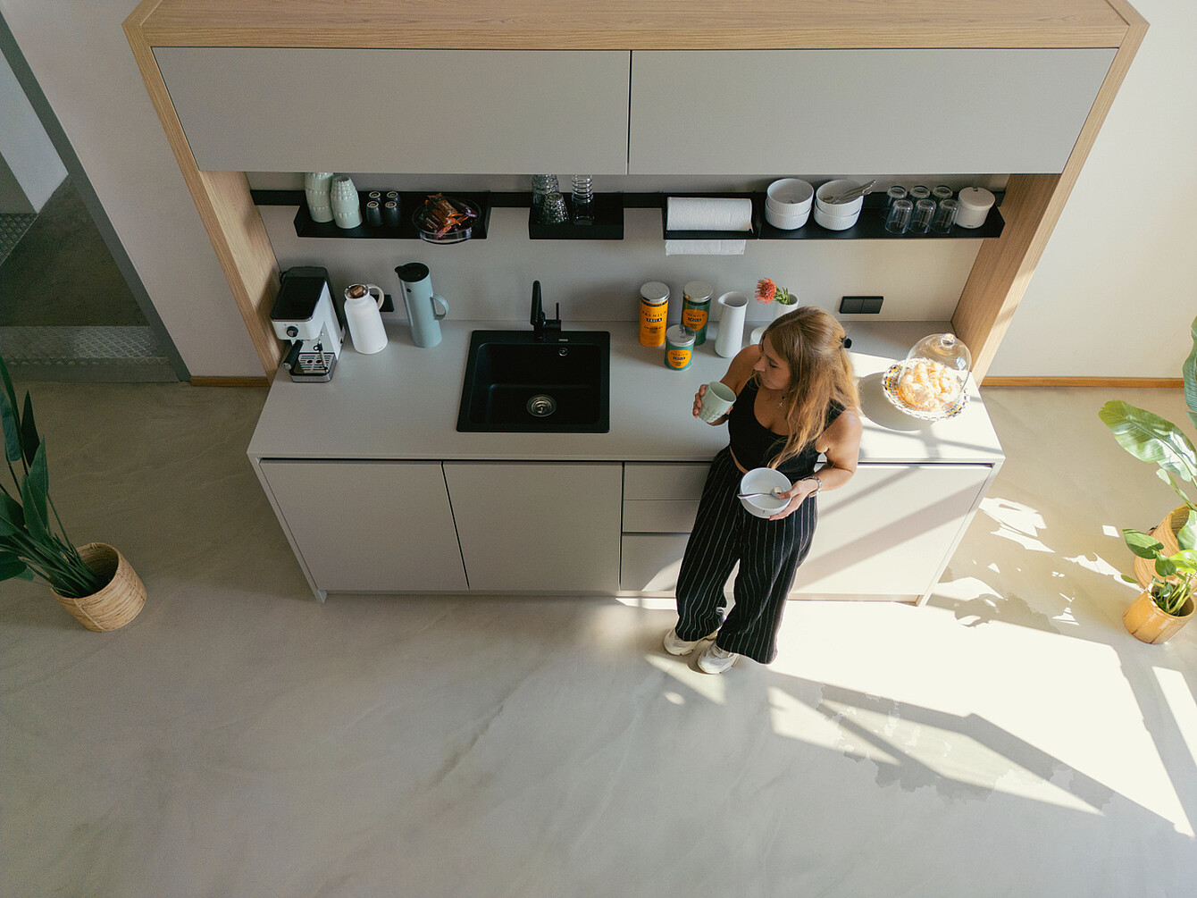 Mujer con top negro y pantalones a rayas de pie en la luminosa cocina de una oficina con luz natural, sosteniendo una taza y un bol cerca de una encimera con una cafetera, tarros y plantas
