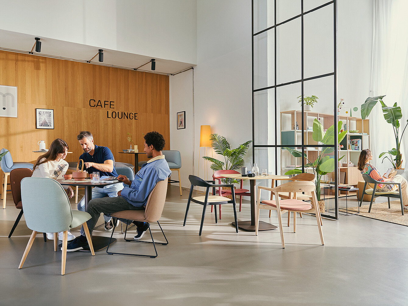 Drei Personen sitzen im modernen Work Café mit Café Lounge Schriftzug an Holztischen, umgeben von Pflanzen und bequemen Stühlen