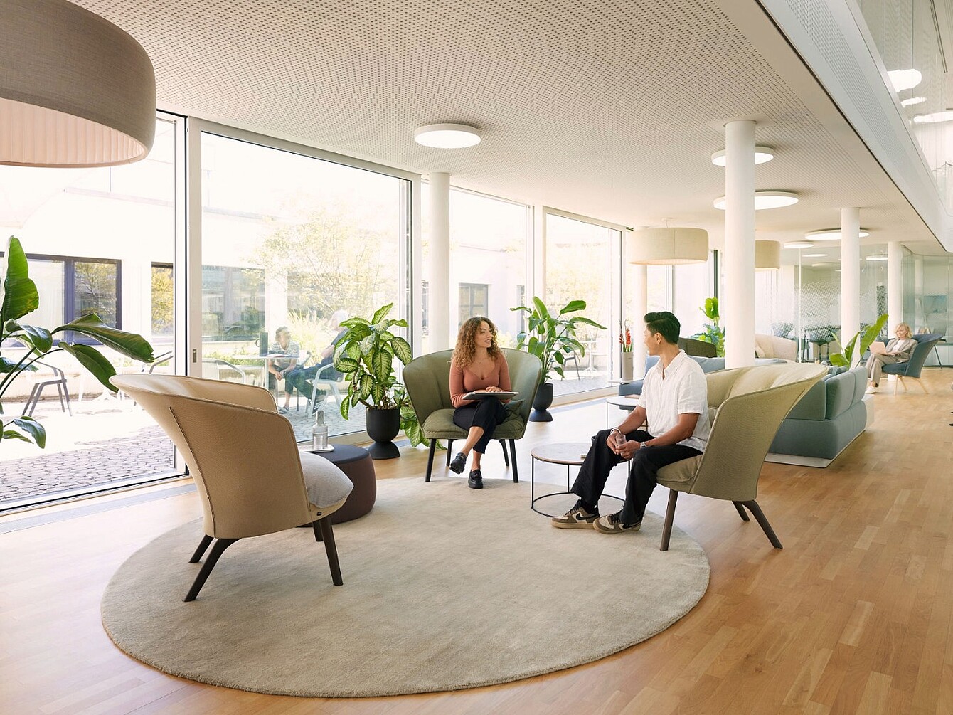 Two people sitting in beige armchairs around a small round table on a circular rug in a spacious, well-lit room with large windows and plants