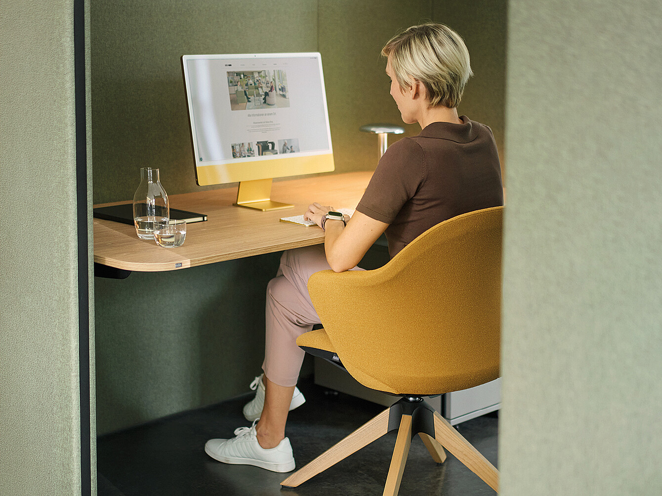 Una persona vestita sportiva è seduta a una scrivania moderna in uno spazio di lavoro separato e insonorizzato. La sedia da scrivania è gialla e ha una forma arrotondata. Sulla scrivania ci sono un grande computer con un supporto giallo, un bicchiere d’acqua e un taccuino. Le pareti dello spazio di lavoro sono rivestite con un tessuto verdastro, che crea un’atmosfera tranquilla.