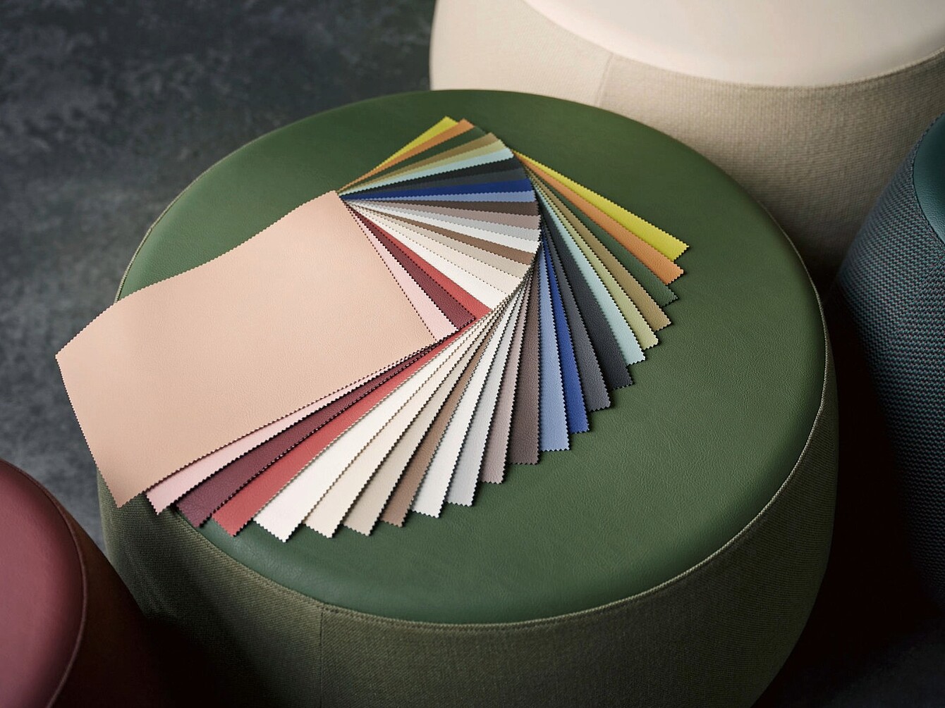 Abanico de muestras de tela de varios colores dispuestas sobre una otomana redonda verde, con partes de otras otomanas visibles a su alrededor