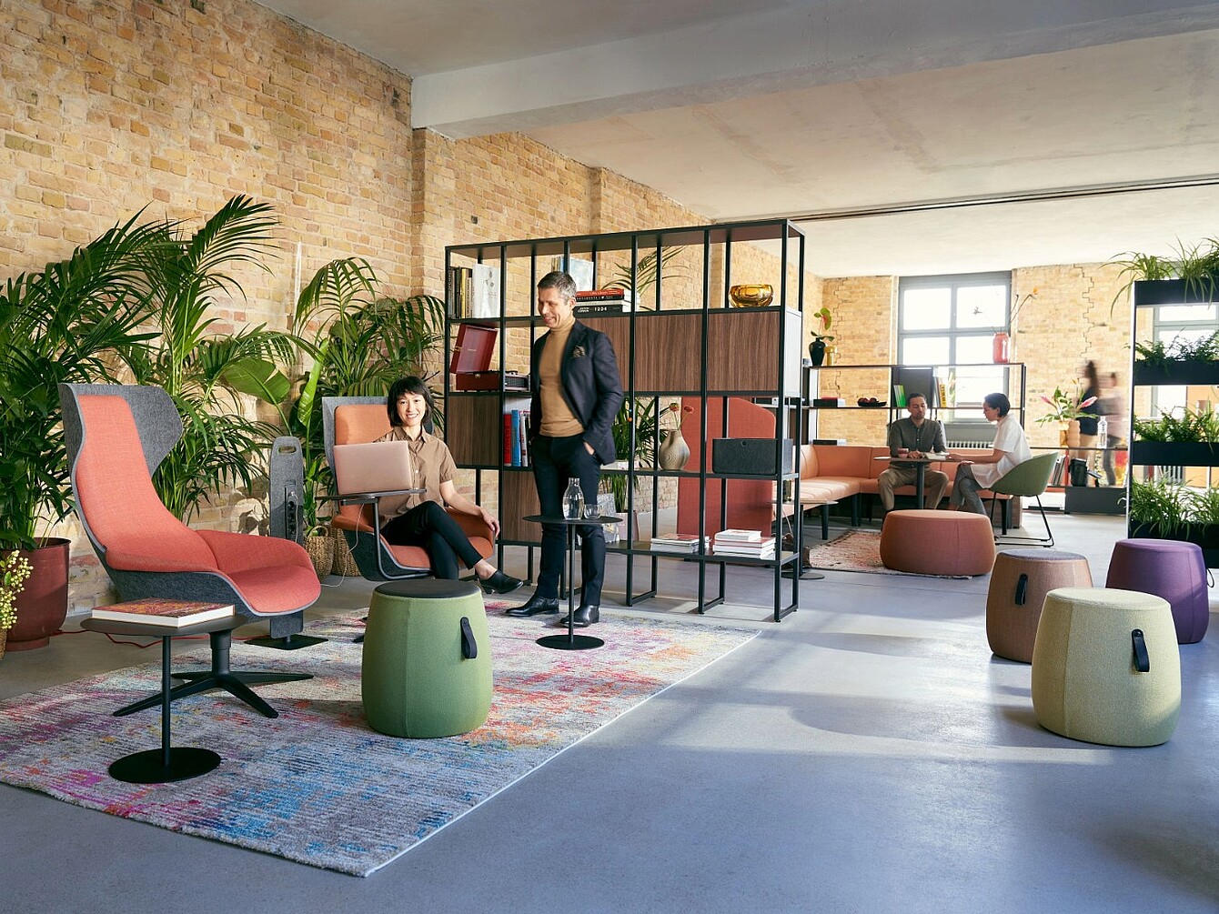Area lounge aperta con sedute moderne tra cui poltrone, sgabelli e un divano modulare separati da una scaffalatura in metallo nero e legno, sullo sfondo di una parete in mattoni con grandi finestre e piante da interno