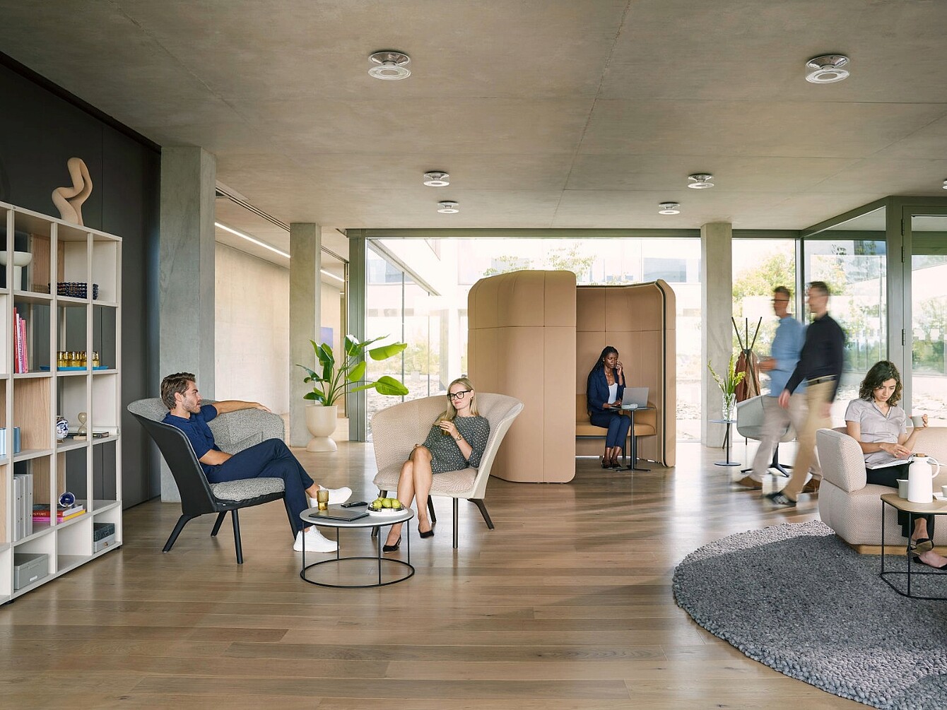Modern interior with seating furniture, bookshelf, plants, and two people in a partitioned seating nook, other people sitting or walking in the space