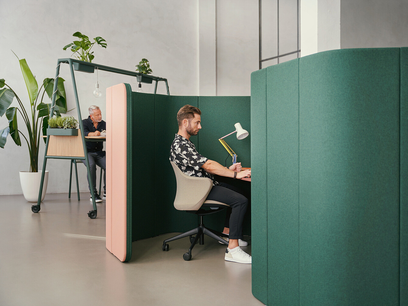 Hombre trabajando en una cabina acústica ecológica en una oficina moderna con luz natural y plantas, que refleja las prioridades de bienestar en el lugar de trabajo.