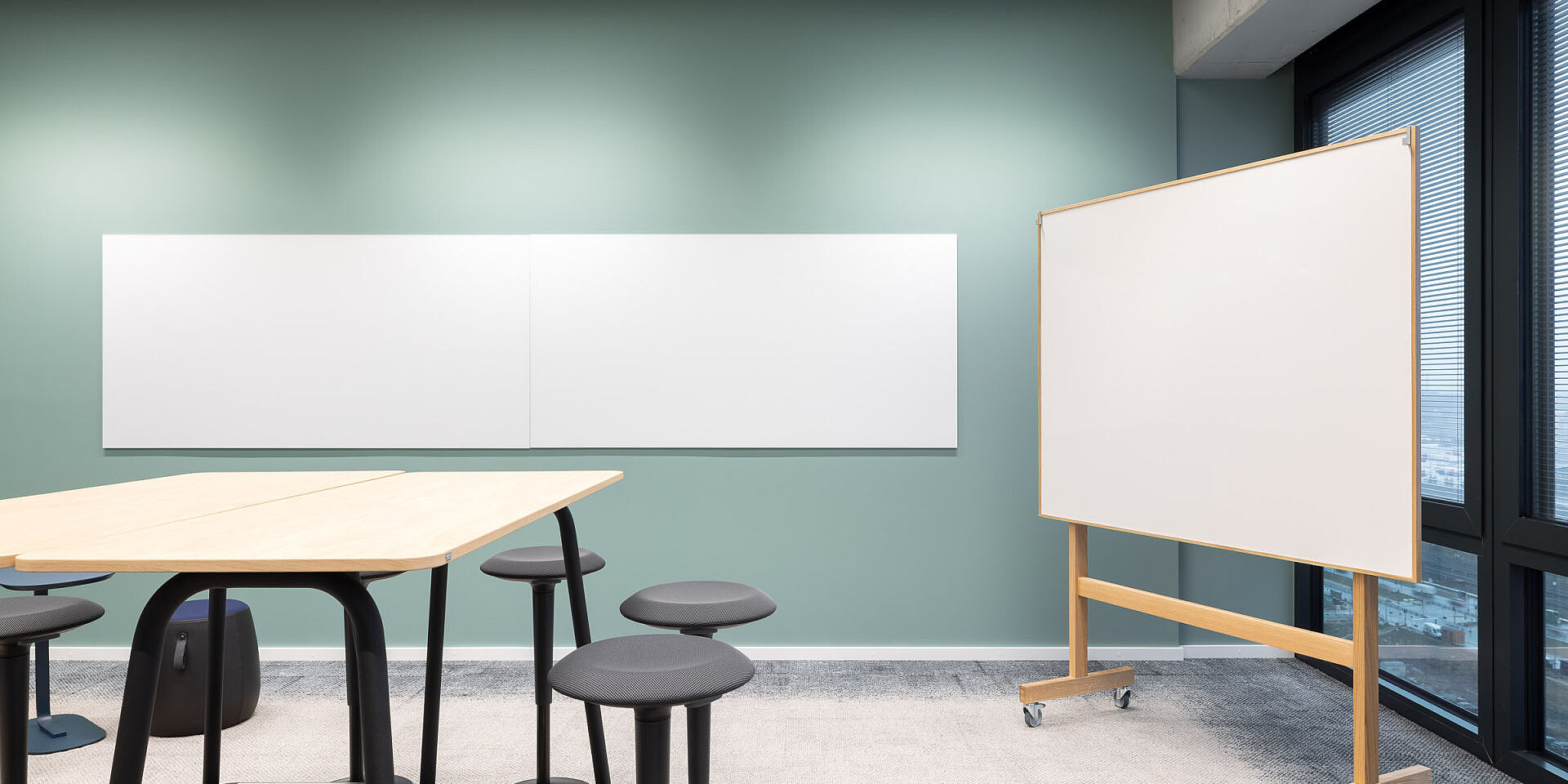 Modern workspace with se:lab high desk workshop table, se:fit standing seat and several universal se:dot stools in front of a green wall and mobile whiteboard. 
