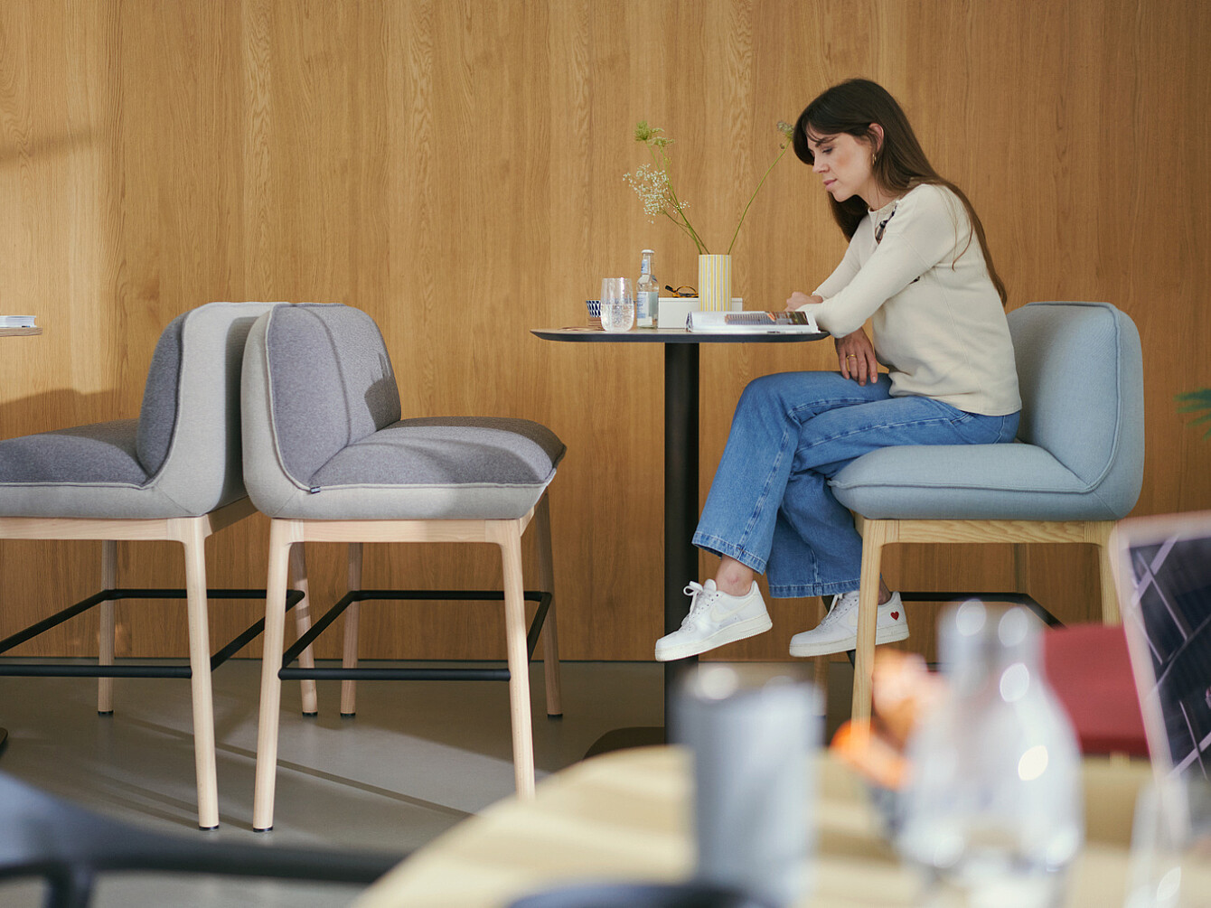 Mujer sentada sola en una pequeña mesa en una moderna sala de descanso con paredes de madera, leyendo o trabajando en silencio, lo que ilustra un entorno tranquilo y propicio para el bienestar de los empleados.