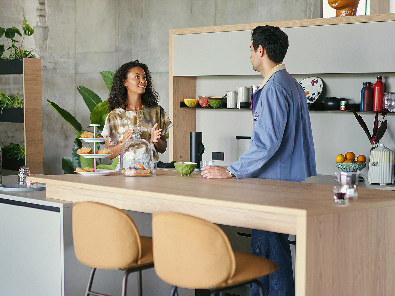 Due dipendenti che conversano a un bancone in legno in un work café con piante e oggetti da cucina intorno, a illustrare un ambiente di lavoro flessibile.