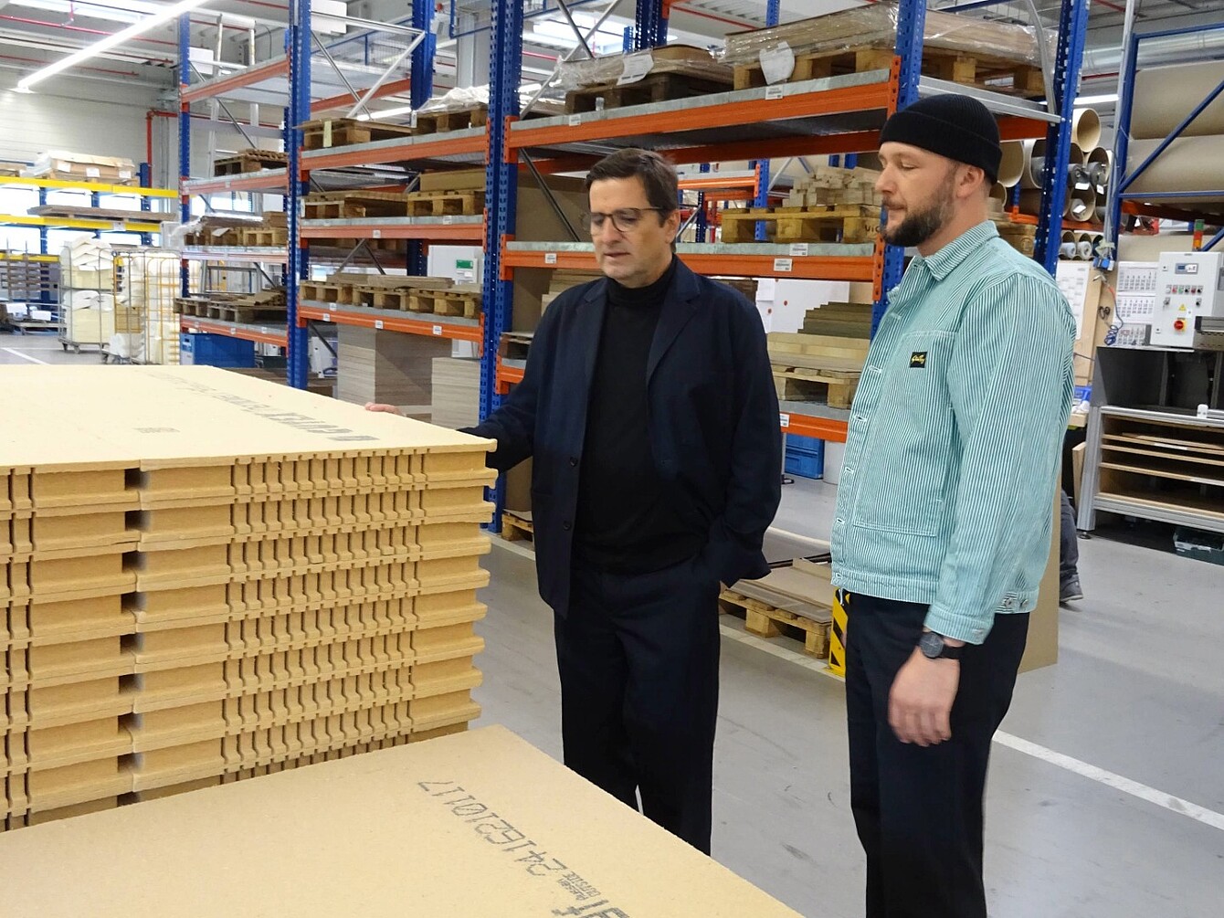 Dos hombres (Rudi Raschke y Konstantin Thomas) están junto a pilas de paneles de fibra de madera en un almacén con estanterías metálicas y equipo industrial.