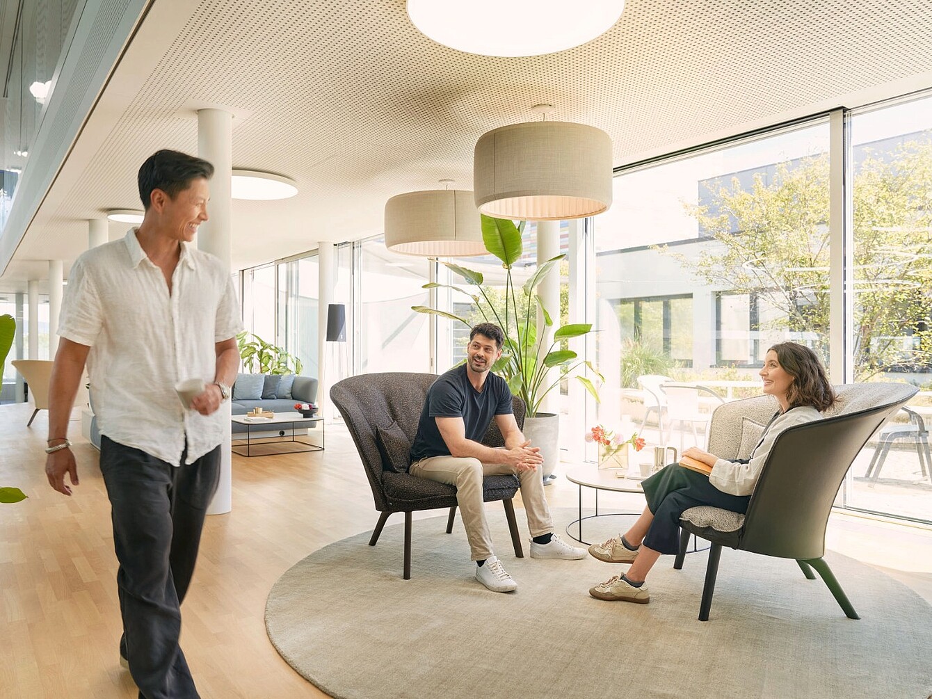 Drie mensen in een moderne kantoorlounge met grote ramen, zittend en staand rond een rond tapijt en fauteuils