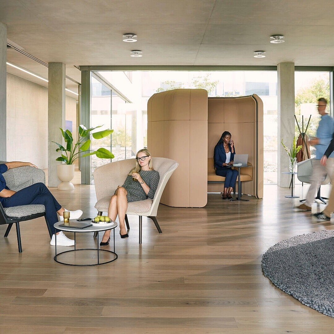 Open office space with soft seating including armchairs and a curved partition creating semi-private zones where people are sitting, working, and conversing