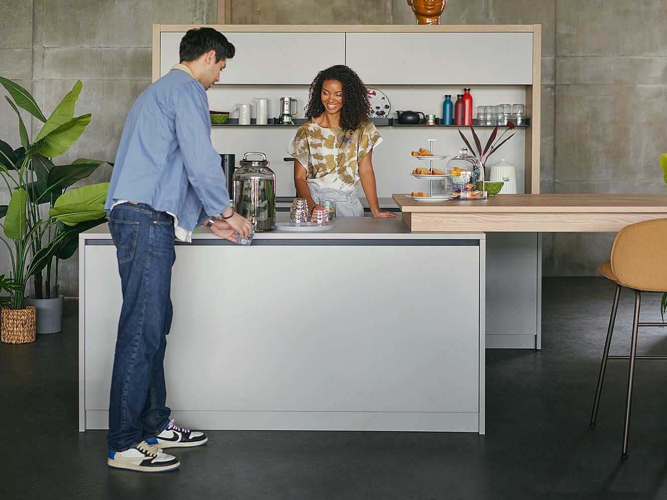 Due persone interagiscono nella zona cucina di uno spazio di lavoro ibrido e duttile moderno con piante, snack e bevande, che enfatizza la flessibilità e la comunità.