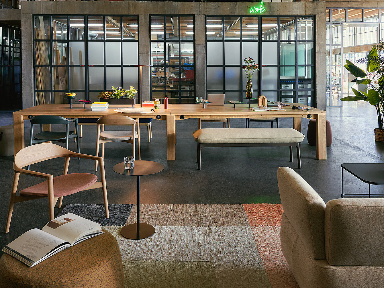 Modernes Büro mit großen Holztischen, Stühlen, einer Bank und Pflanzen in einem Raum mit Glaswänden und Betonboden