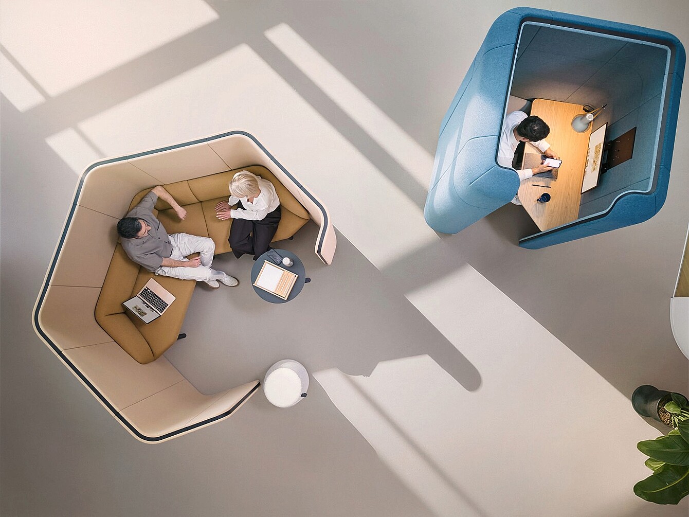Dos personas sentadas en un refugio textil beige con un ordenador portátil y una mesita, y una persona trabajando sola en un refugio textil azul con un escritorio y una lámpara, todo ello visto desde arriba en una habitación luminosa con sombras y una planta visible.