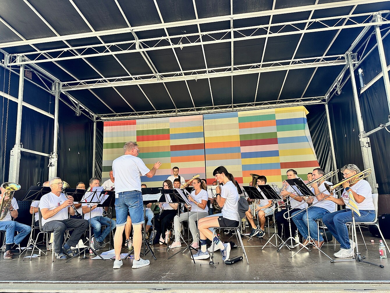 Dirigent der Sedus Werksmusik in weißem T-Shirt und Jeansshorts leitet ein Orchester mit Blasinstrumenten auf einer Bühne mit buntem, gestreiftem Hintergrund.