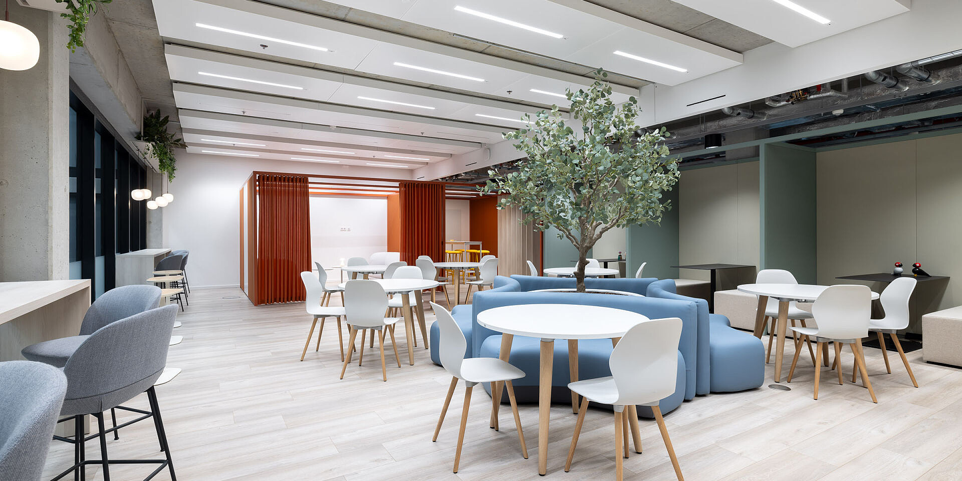 Modern bistro area at Veolia with round se:assist tables and ergonomic se:spot designer chairs in light colours, surrounded by sofas and a large plant for relaxed breaks. 