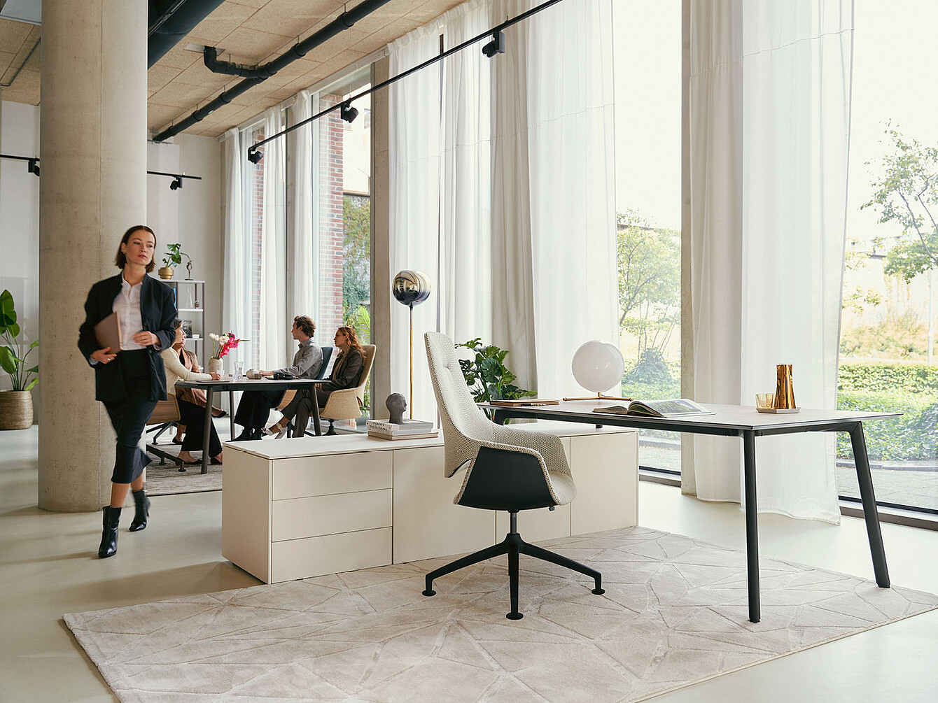 Espacio de oficina moderno con grandes ventanas, cortinas blancas, un escritorio con una silla giratoria Sedus se:mission blanca y personas sentadas y caminando