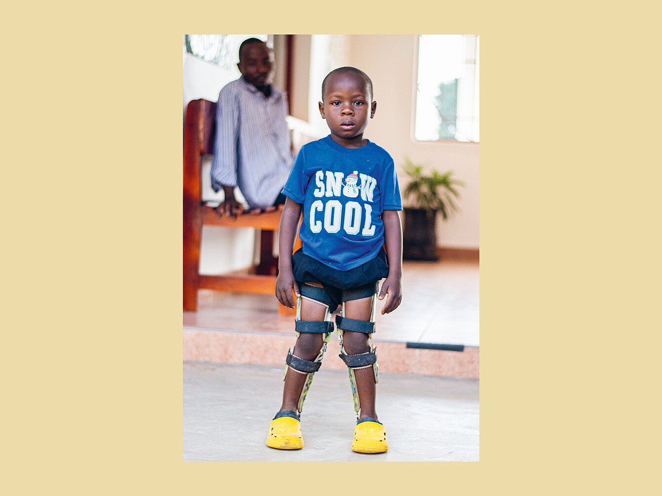 Niño de pie en interior con camiseta azul con el texto «SNOW COOL» y un gráfico de un muñeco de nieve, pantalones cortos negros, aparatos ortopédicos en las piernas y zuecos amarillos.