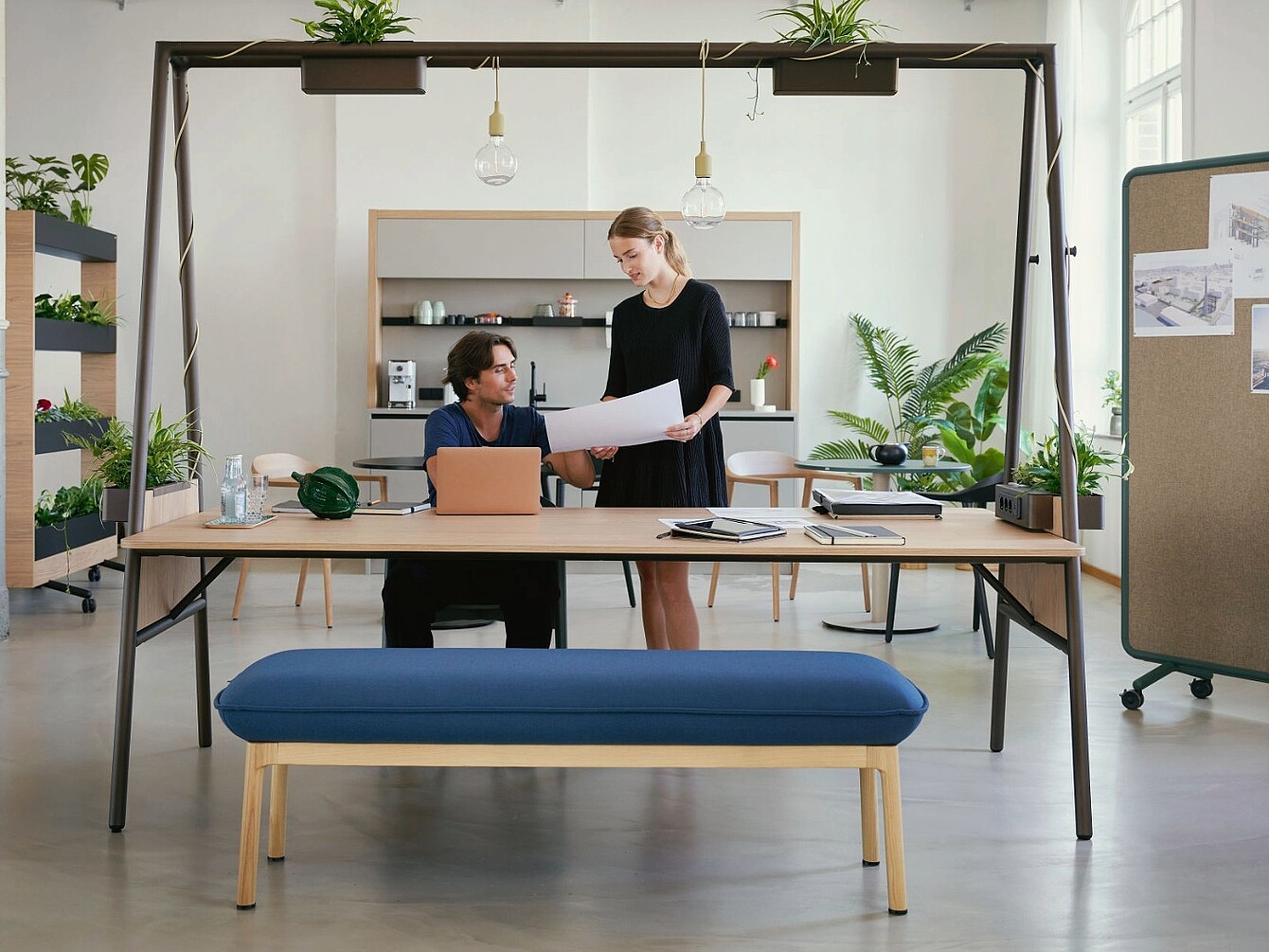 De afbeelding toont een moderne werkplek met een groot bureau waar twee mensen samen documenten bekijken. Boven het bureau bevindt zich een open frameconstructie met geïntegreerde planten en verlichting, die de werkruimte visueel afbakent. 