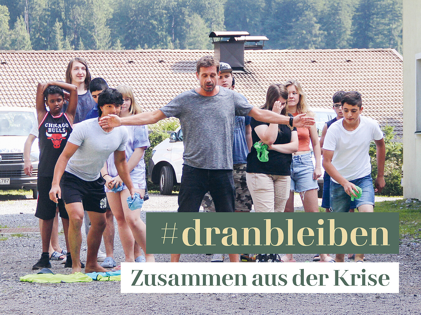 Jugendliche stehen im Freien auf einer Wiese vor einem Haus, einige in sportlicher Kleidung, eine trägt ein Chicago Bulls T-Shirt, sie bereiten sich auf eine Aktivität vor.