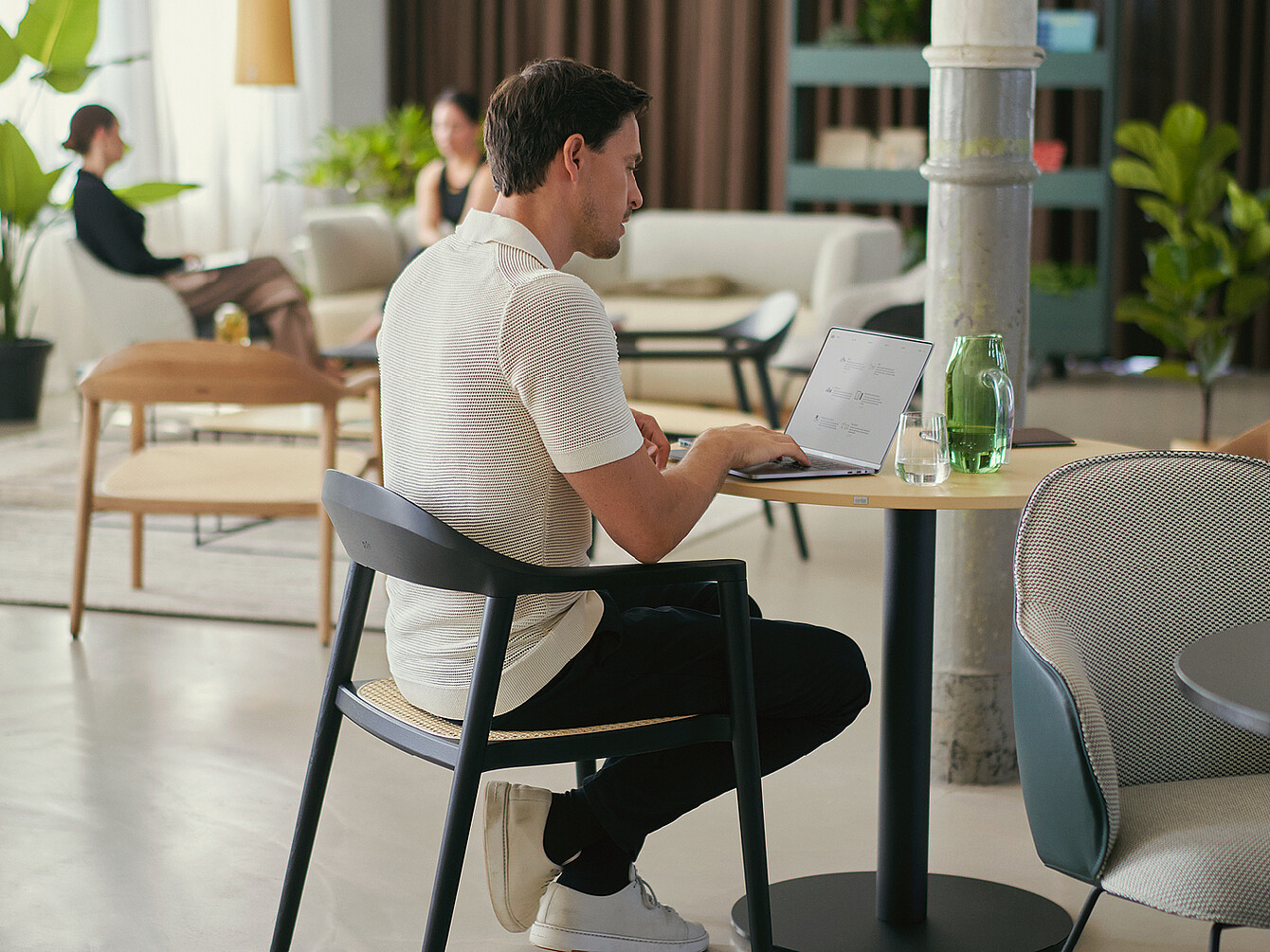 Persona seduta a un tavolo rotondo in un work café, che utilizza un laptop con un bicchiere d’acqua e una bottiglia verde accanto, a illustrazione delle atmosfere variegate che stimolano il coinvolgimento dei dipendenti.