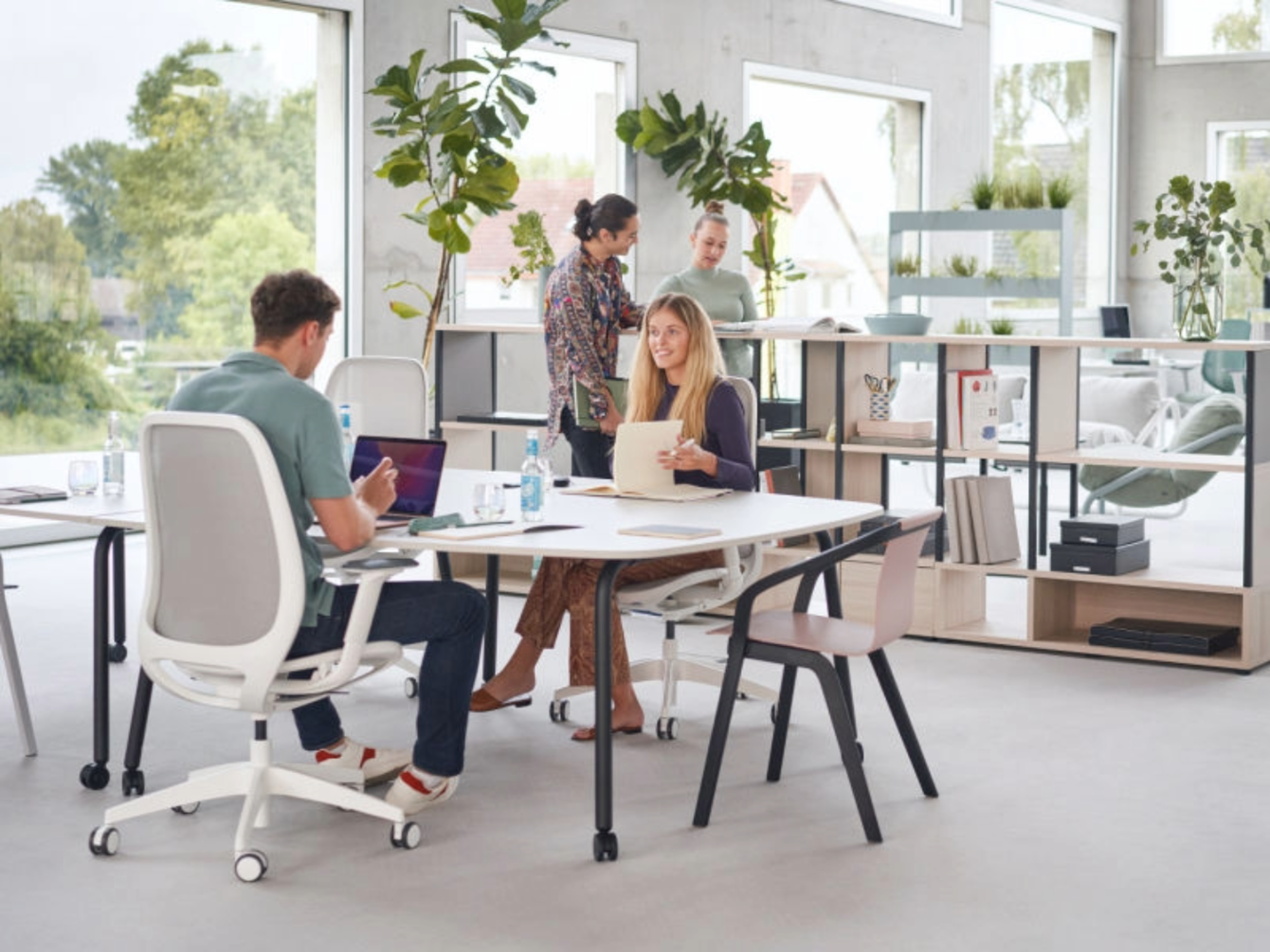 Vier Personen sitzen und stehen an einem hellen Tisch in einem modernen Büro mit großen Fenstern und Pflanzen